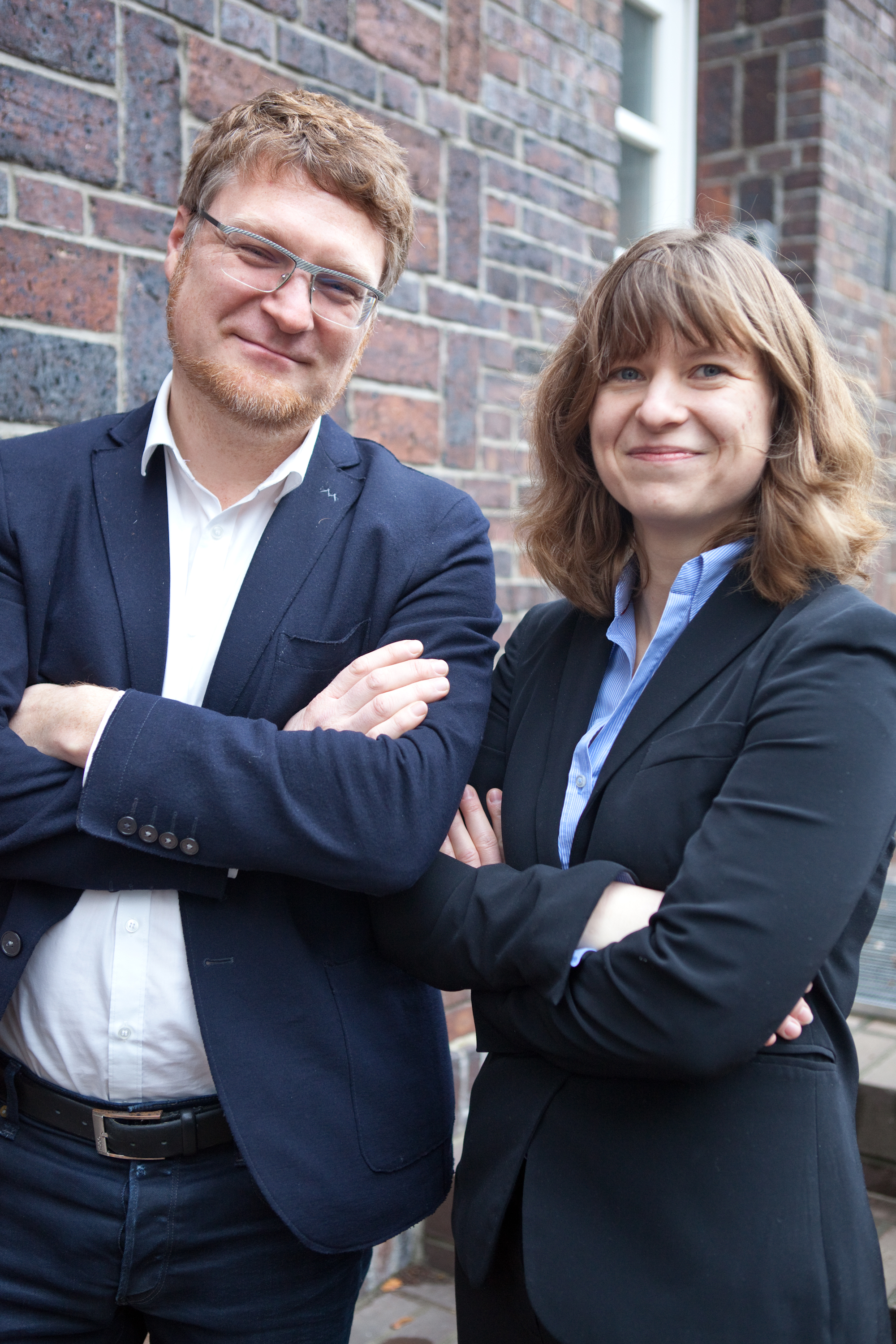 Zwei Personen in Business-Outfits vor Backsteinwand, Arme verschränkt – Teamfoto draußen.
