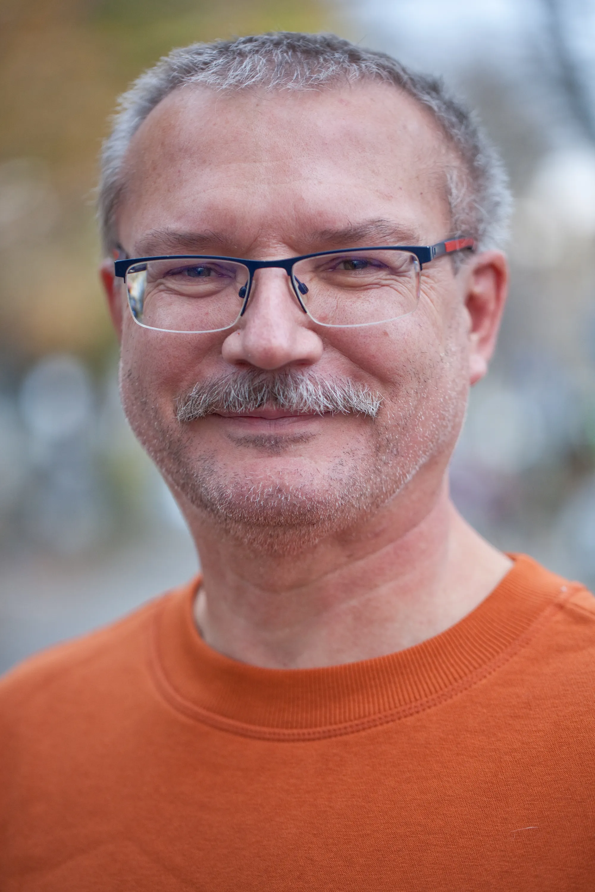 Porträt eines älteren Mannes mit Brille, orangefarbenes Oberteil, Bäume im Hintergrund – Mitarbeiter.