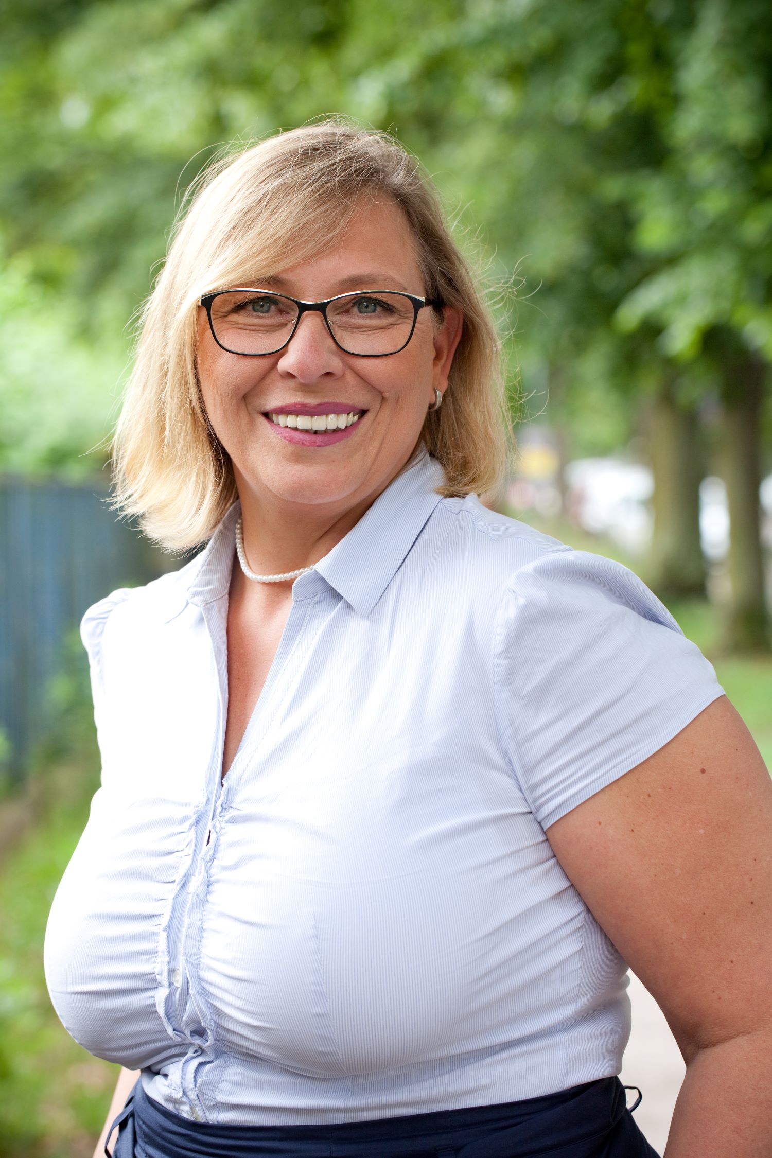 Porträt einer Frau mit blonder schulterlangen Haaren und Brille, weißes Oberteil, Naturhintergrund