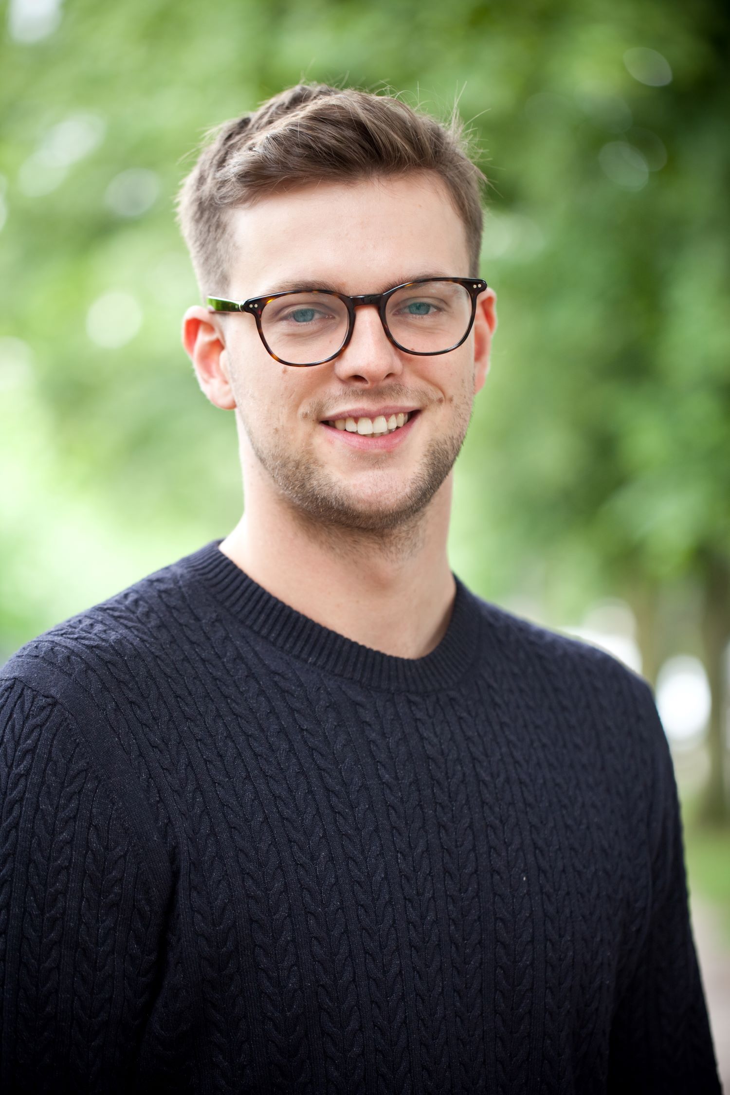 Porträt eines jungen Mannes mit Brille und Bart, lächelnd, Bäume im Hintergrund.