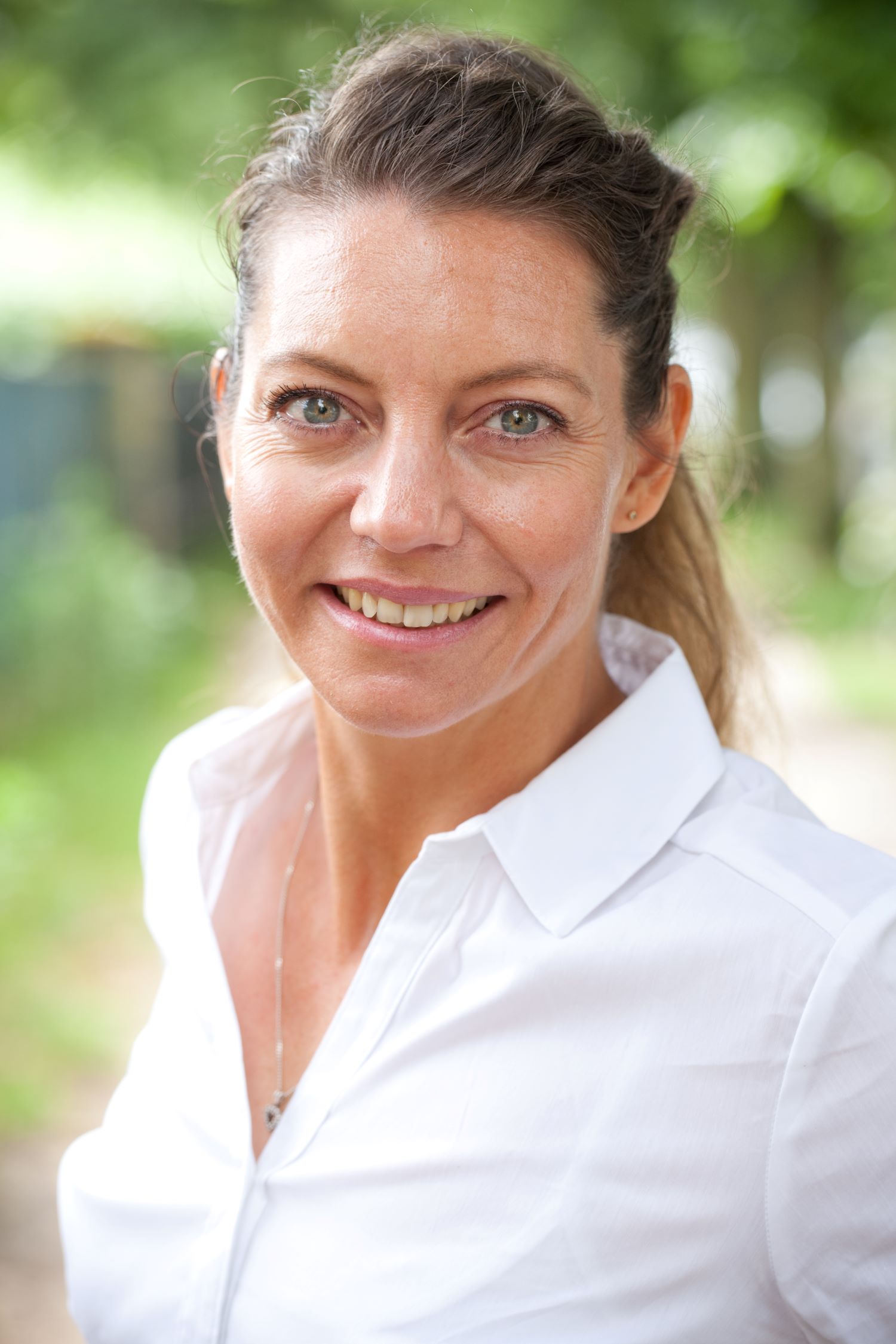 Porträt einer Frau mit zusammengebundenen Haaren, weißer Bluse, Natur im Hintergrund.