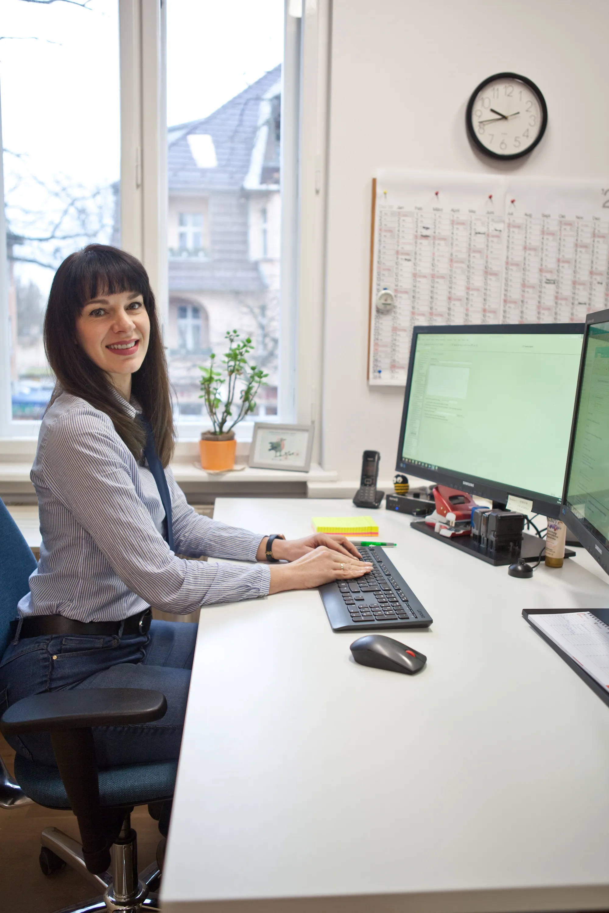 Frau sitzt am Schreibtisch mit zwei Monitoren, Blick zur Kamera – Arbeitsplatz.

