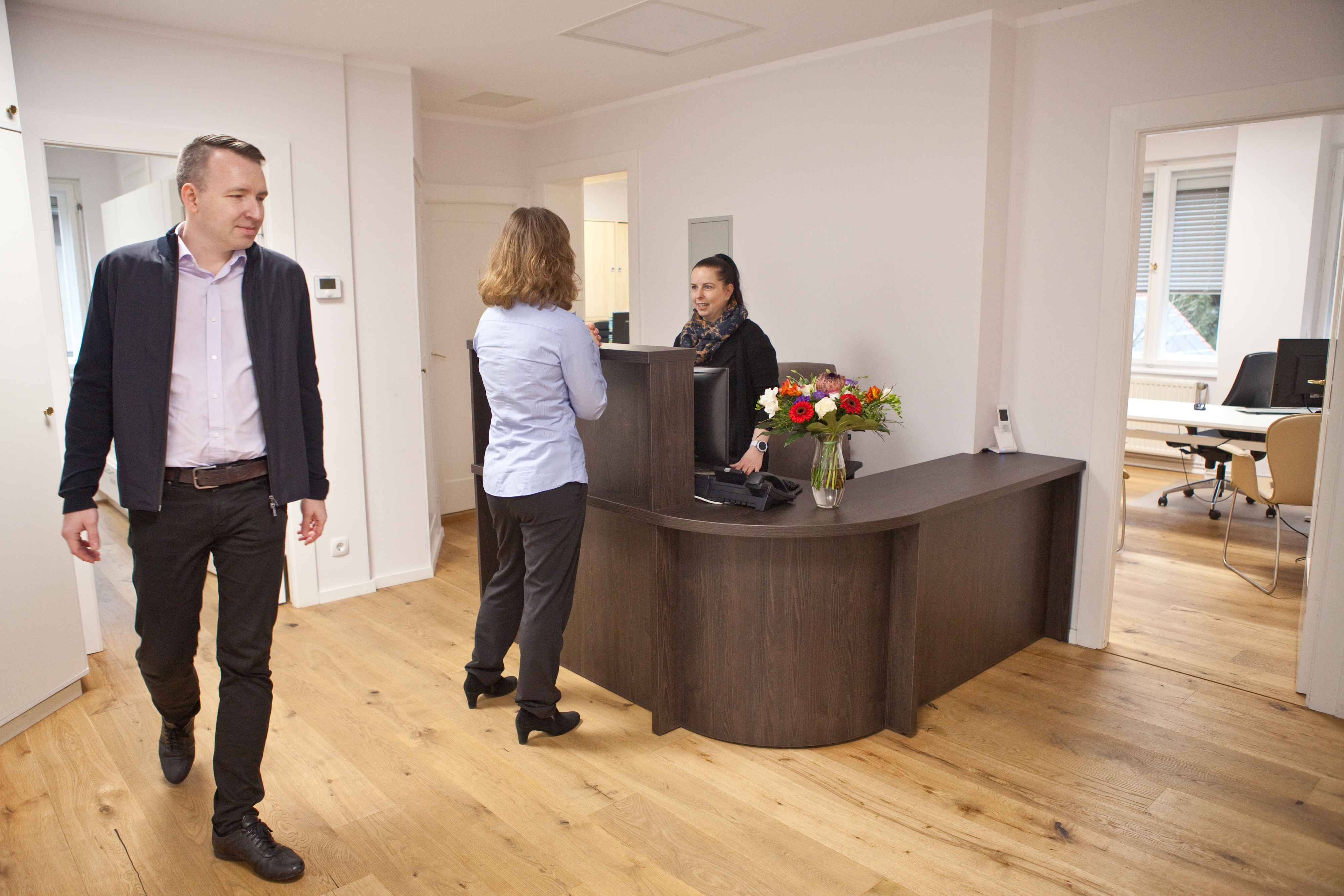 Zwei Männer gehen durch Flur, Empfangstresen im Hintergrund – Büroalltag.